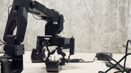 A robotic arm fitted with a 3D-printed compliant flexure gripper picking up a small component on a workbench